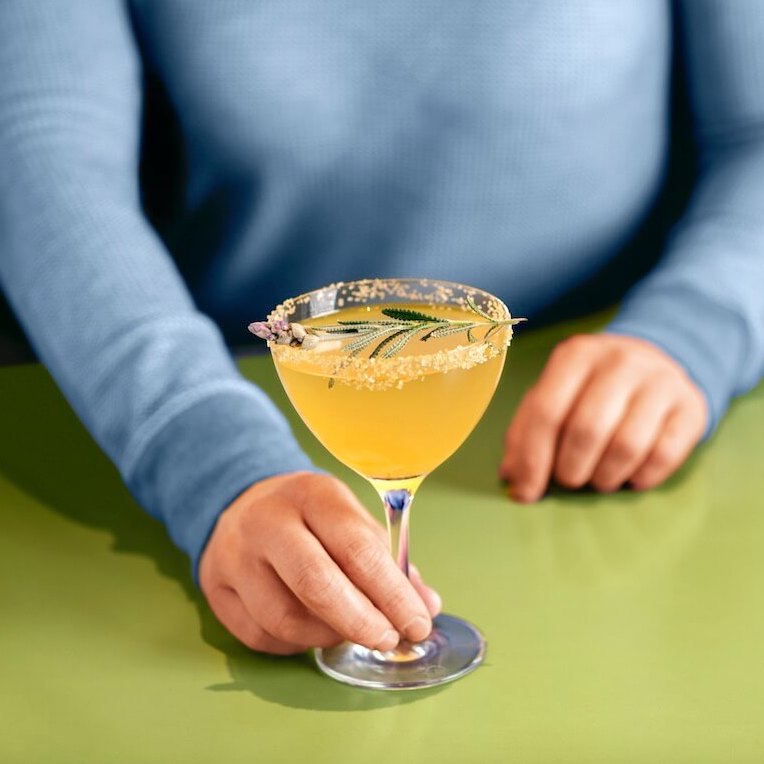 Woman in blue sweater holding the Snooze Abri-ca-lav-ra Mimosa; a combination of Snooze Sparkling, Apricot Liqueur, Fresh Lavender, Honey and Lemon Juice
